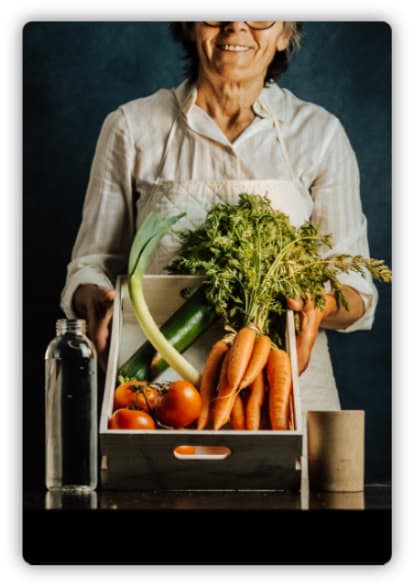 An old woman presenting a basket of vegetables including carrots, tomatoes, and zucchini