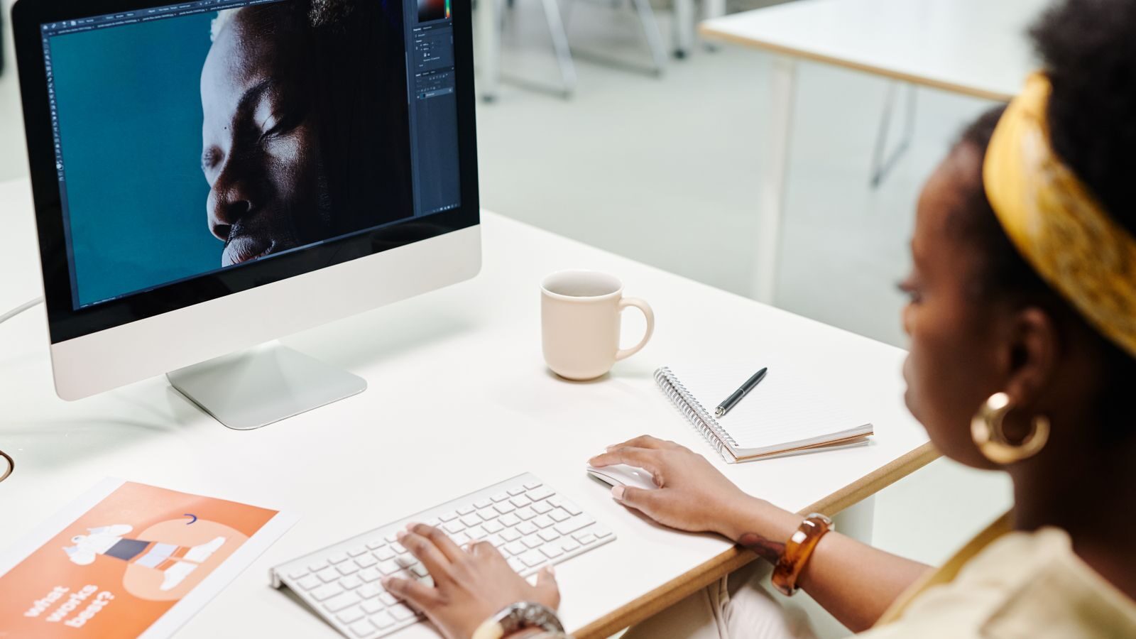 a designer at a bright white desk in an open-plan office, reviewing an image on a large monitor