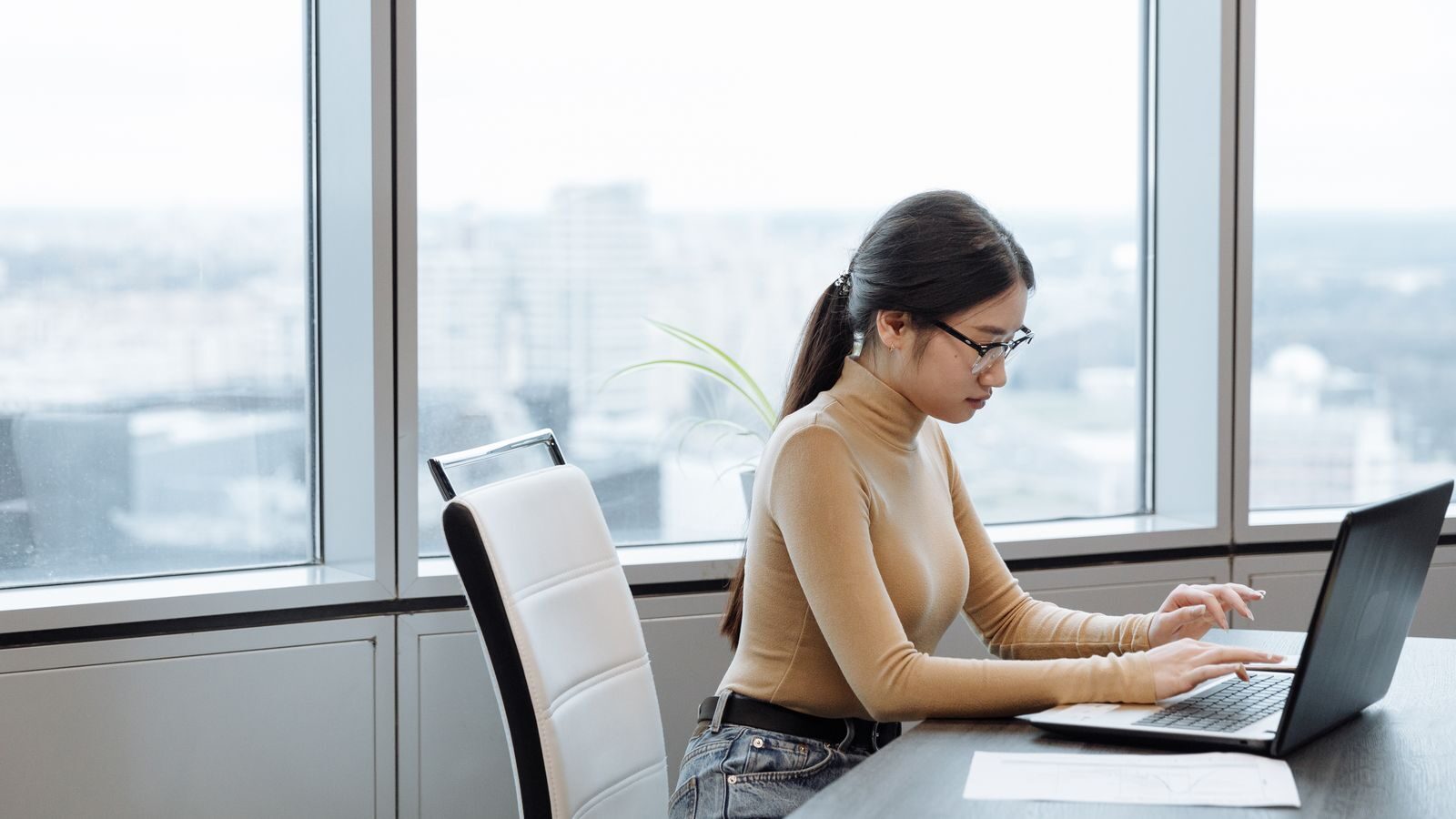 a person typing on a laptop at a desk beside floor-to-ceiling office windows overlooking a city